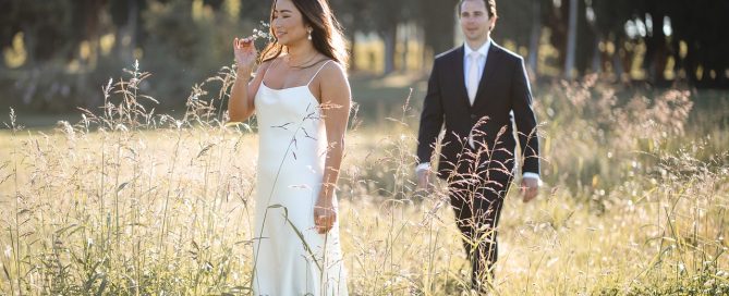 & groom standing in a lush, outdoor setting with a pastel sunset sky, a hand-crafted archway draped with foliage, and a creative seating arrangement of colorful chairs