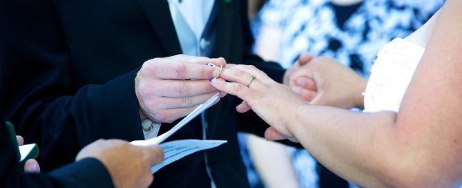 -up of a couple holding hands, each wearing a wedding band, with a background of traditional elements like a bouquet of flowers, an arch of lace, and a veil