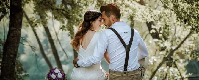 E of a bride and groom smiling and embracing in front of an officiant wearing a long white robe and holding a golden book