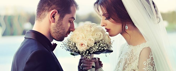 G couple in a garden, holding hands and smiling tenderly at each other, with a beautiful white rose bouquet
